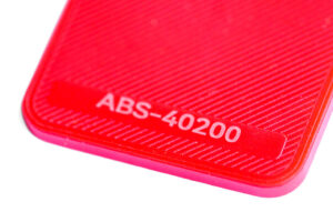 ABS 40200 - KIT3D GmbH – 3D-Technologie & Konstruktion Close-up of a red plastic surface with diagonal texture and the label ABS-40200 on it.