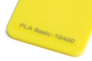 PLA Basic 10400 - KIT3D GmbH – 3D-Technologie & Konstruktion Close-up of a bright yellow plastic object with the text 'PLA Basic-10400' along its edge, showing a textured corner