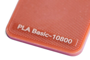 PLA Basic 10800 - KIT3D GmbH – 3D-Technologie & Konstruktion Close-up of an orange PLA Basic-10800 label on a textured surface of a 3D printing filament package, with diagonal grooves.