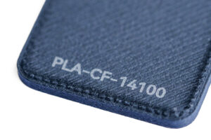PLA CF 14100 - KIT3D GmbH – 3D-Technologie & Konstruktion Close-up of a dark blue textured surface labeled PLA-CF-14100, likely a 3D printer filament sample.