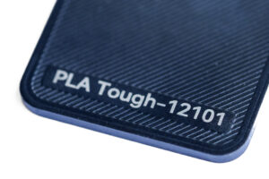 PLA Tough 12101 - KIT3D GmbH – 3D-Technologie & Konstruktion Close-up of a dark blue textured surface showing the label PLA Tough-12101.