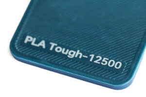 PLA Tough 12500 - KIT3D GmbH – 3D-Technologie & Konstruktion Close-up of a teal textured surface with embossed text: 'PLA Tough-12500'.